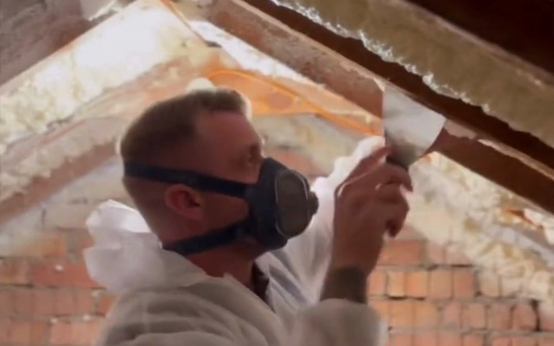 A remover in a white protective suit and mask scraping spray foam off the rafters